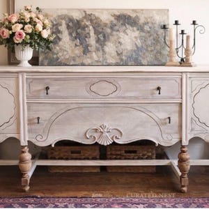 Könnte beinhalten: Ein weißes Sideboard im Used-Look mit verzierten Details, zwei Schubladen und Schranktüren mit dekorativen Ausschnitten. Das Sideboard hat ein unteres Regal mit zwei geflochtenen Körben und ist mit einem Blumenarrangement und Kunstwerk gekrönt.