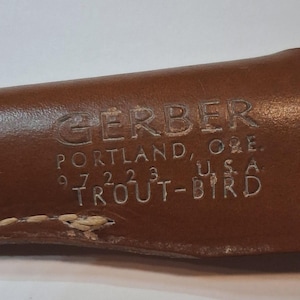以下が含まれることがあります： 「GERBER PORTLAND, ORE. 97223 TROUT-BIRD U.S.A.」のエンボス加工が施された茶色の革製シースのクローズアップ。シースの端にはステッチが見えます。