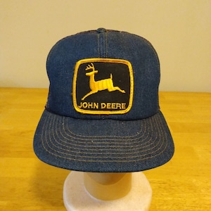 Puede incluir: Una gorra de camionero de mezclilla azul con un logotipo de John Deere amarillo que presenta un ciervo saltando. El logotipo está en un parche negro y amarillo.