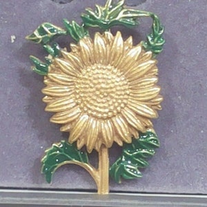 Könnte beinhalten: Eine goldfarbene Sonnenblumenbrosche mit grünen Emailleblättern. Die Sonnenblume hat eine detaillierte Mitte und Blütenblätter.