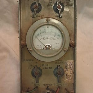 May include: Vintage metal multimeter with multiple dials, knobs, and a central gauge. Features settings for voltage, ohms, and battery testing. The device has a worn, industrial aesthetic with labels and text.