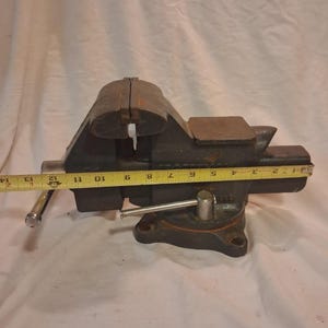 May include: A heavy-duty black metal vise with a silver handle and a yellow measuring tape showing a length of approximately 13 inches. The vise has a sturdy base with bolt holes.