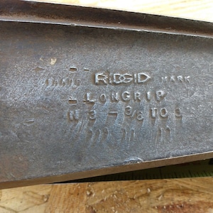 May include: Close-up of a metal tool with the text "RIDGID MARK LONG-RIP IN 3 7/8 TO 10" engraved on the surface.