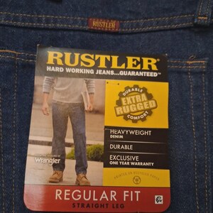 May include: Close-up of blue denim jeans with a tag. The tag reads "RUSTLER" and "HARD WORKING JEANS...GUARANTEED." Features include "DURABLE EXTRA RUGGED COMFORT," "HEAVYWEIGHT DENIM," and "REGULAR FIT STRAIGHT LEG."