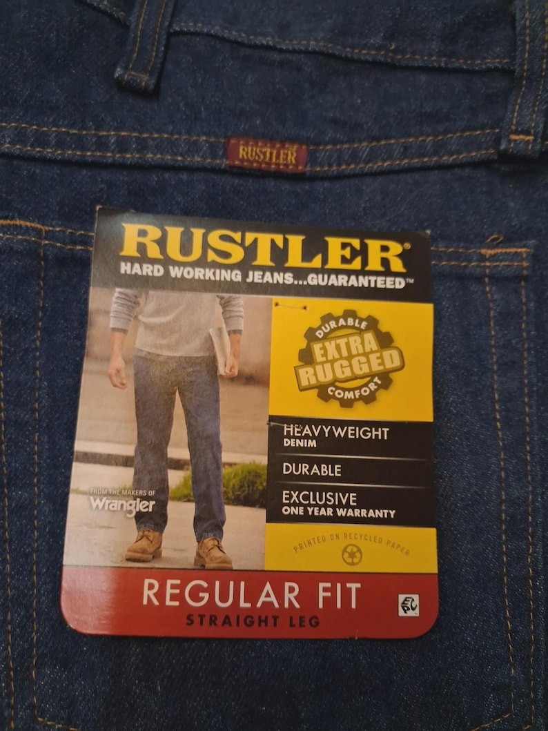 May include: Close-up of blue denim jeans with a tag. The tag reads "RUSTLER" and "HARD WORKING JEANS...GUARANTEED." Features include "DURABLE EXTRA RUGGED COMFORT," "HEAVYWEIGHT DENIM," and "REGULAR FIT STRAIGHT LEG."