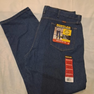 May include: Dark blue denim jeans with the "RUSTLER" brand label visible on the back pocket. The jeans are a regular fit and have a tag with size information. The jeans are folded and laid flat on a white surface.