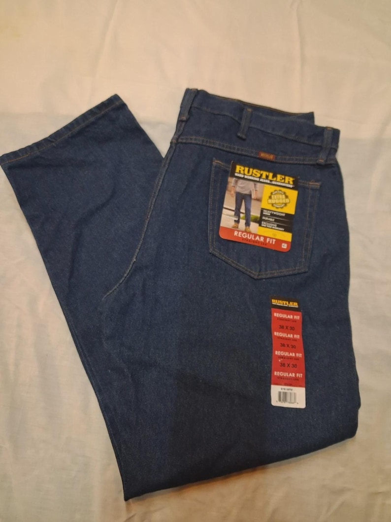 May include: Dark blue denim jeans with the "RUSTLER" brand label visible on the back pocket. The jeans are a regular fit and have a tag with size information. The jeans are folded and laid flat on a white surface.