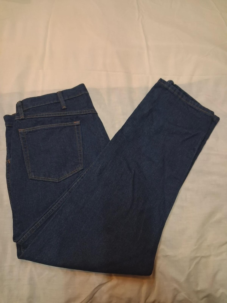 May include: Dark blue denim jeans with a classic five-pocket design. The jeans are folded and laid flat, showing the back pockets and the straight leg cut. The fabric appears to be a medium-weight denim.