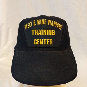 May include: Black baseball cap with a yellow embroidered patch that reads "Fleet & Mine Warfare Training Centre".