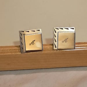 Puede incluir: Un par de gemelos cuadrados con centro dorado y borde plateado. Cada gemelo presenta la letra "A" en una escritura elegante. El borde tiene un diseño geométrico.