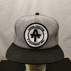 Op de afbeelding: Een grijze en zwarte baseballpet met een witte patch waarop "Akron Tractor & Equipment Inc." staat, met een gestileerde "A" in het midden.
