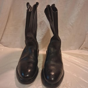Könnte beinhalten: Ein Paar wadenhohe Stiefel aus schwarzem Leder mit abgerundeter Zehenpartie, glatter Oberfläche und Anziehlaschen am Schaft. Die Stiefel haben eine robuste Konstruktion mit sichtbaren Nähten an der Sohle und einem niedrigen Blockabsatz.