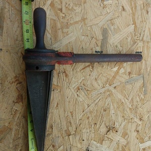 May include: A rusty metal pipe threading tool with a wooden handle and a measuring tape in the background.