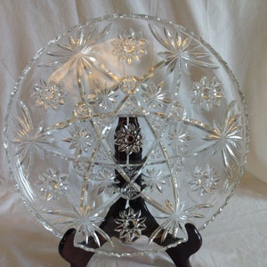 Puede incluir: Un plato de servir grande de vidrio transparente con un patrón de estrella. El plato está sobre un soporte de madera.