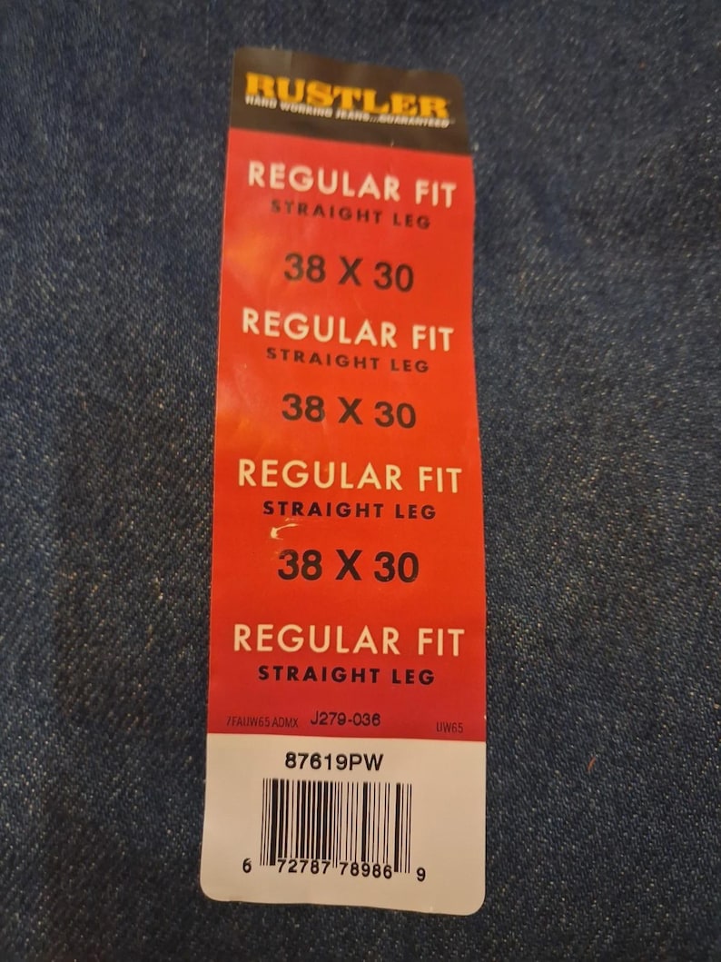 May include: A red tag for Rustler jeans, with the text "REGULAR FIT STRAIGHT LEG" and size "38 X 30". The tag also includes a barcode and the text "87619PW". The tag is on a blue denim background.