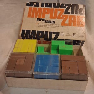 May include: A vintage Impuzzables three-dimensional perceptual puzzle set in a cardboard box. The set includes multiple clear plastic containers holding yellow, green, brown, and blue geometric plastic blocks for solving various puzzles.