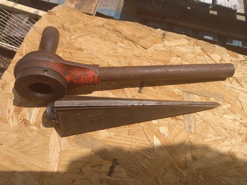 May include: A rusty, red and black pipe wrench with a long handle and a wedge-shaped tool. The tools are on a wooden surface.