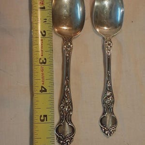 Puede incluir: Dos cucharas de plata con diseños florales y de volutas ornamentados en los mangos. Las cucharas miden aproximadamente 15 cm de largo. Una cinta métrica amarilla está colocada junto a las cucharas.