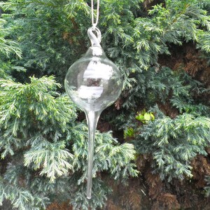 May include: A clear glass ornament with a long, pointed tip. The ornament is hanging from a string and is suspended in front of a green bush.