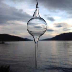 May include: A clear glass ornament shaped like a teardrop with a pointed tip. The ornament is hanging from a string and is reflecting the sky and water in the background.