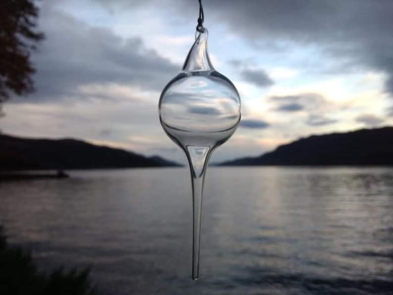 May include: A clear glass ornament shaped like a teardrop with a pointed tip. The ornament is hanging from a string and is reflecting the sky and water in the background.