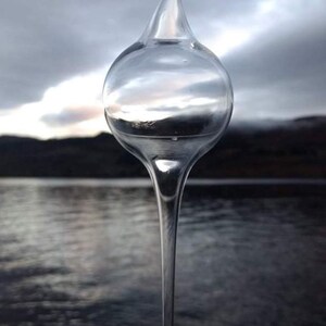 May include: A clear glass ornament with a long, pointed tip hanging against a blurred background of a lake and mountains.