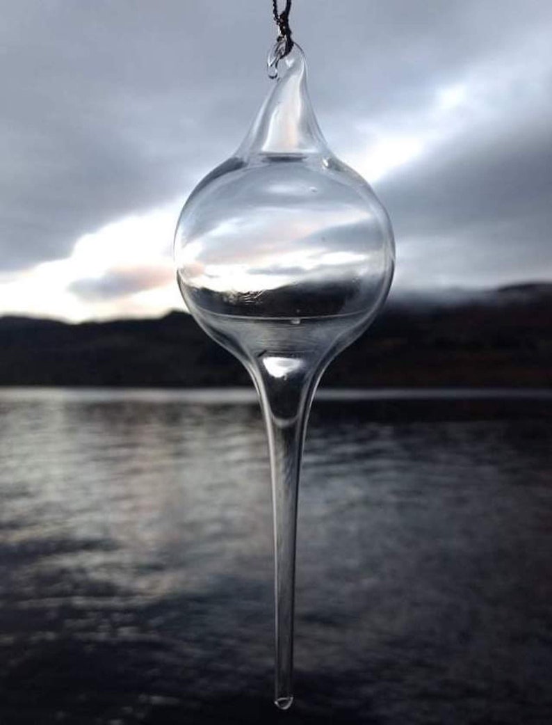 May include: A clear glass ornament with a long, pointed tip hanging against a blurred background of a lake and mountains.