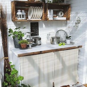 May include: A white kitchen counter with a wooden top and two white fabric curtains. The counter has a variety of items on it, including plants, dishes, and a chalkboard sign. The counter is in a white room with a brick wall.