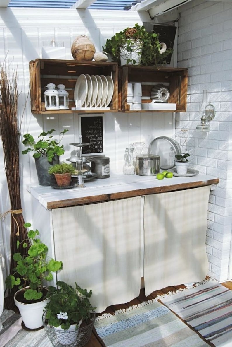 May include: A white kitchen counter with a wooden top and two white fabric curtains. The counter has a variety of items on it, including plants, dishes, and a chalkboard sign. The counter is in a white room with a brick wall.