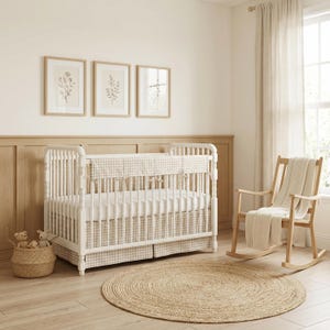 Op de afbeelding: Een kinderkamer met een witte wieg met een gingham patroon bumper en rok. Een gevlochten mand met knuffels staat in de buurt. Een houten schommelstoel met een deken en een rond jute vloerkleed maken de kamer compleet.