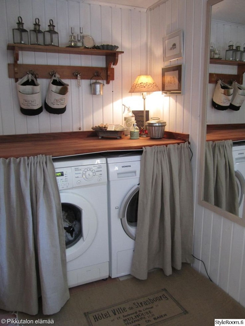 May include: Two white washing machines with a beige curtain covering the front of each machine. The machines are in a white tiled room with a wooden shelf above them. The shelf has a variety of items on it, including a lamp, a mirror, and a few decorative items.