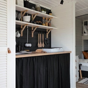 May include: A small kitchen with white walls and black cabinets. The cabinets have shelves with various items, including jars, glasses, and utensils. A black curtain hangs below the counter, covering the lower cabinets. The kitchen has a white countertop with a small stove and a wooden cutting board.