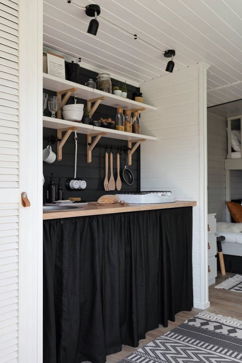 May include: A small kitchen with white walls and black cabinets. The cabinets have shelves with various items, including jars, glasses, and utensils. A black curtain hangs below the counter, covering the lower cabinets. The kitchen has a white countertop with a small stove and a wooden cutting board.