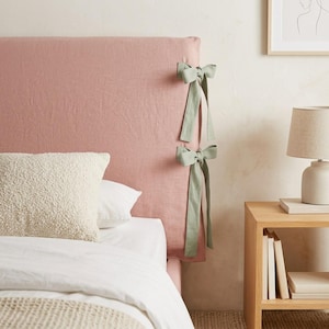 May include: A dusty pink fabric headboard cover featuring two sage green ribbon ties on the side. The cover is draped over a rectangular headboard, with the ties secured in bows. The fabric has a visible woven texture, and the design is simple.