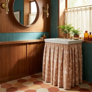 Puede incluir: Un lavabo rectangular blanco montado sobre un marco de madera, oculto por una falda de tela plisada hasta el suelo en color tostado cálido con un estampado floral botánico blanco, situado en una habitación con paneles de madera.