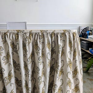 May include: A gathered fabric table skirt featuring a repeating pattern of tigers in shades of gold and dark brown on a light beige background. The fabric is pleated along the top edge and hangs down to cover the front of a rectangular table.