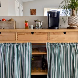 May include: A wooden cabinet with two drawers and two shelves. The cabinet has a green and white striped curtain covering the bottom. A coffee maker and a teapot are on the counter above the cabinet.