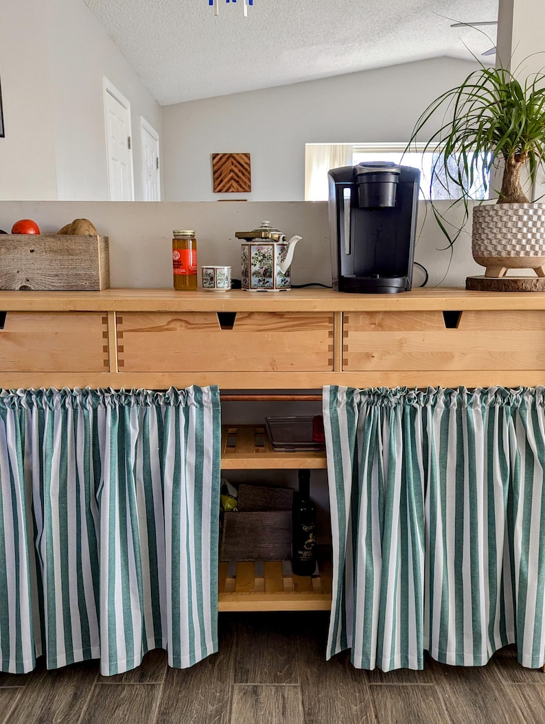 May include: A wooden cabinet with two drawers and two shelves. The cabinet has a green and white striped curtain covering the bottom. A coffee maker and a teapot are on the counter above the cabinet.