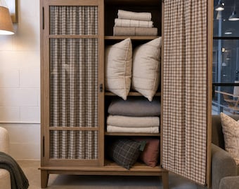 May include: A tall wooden cabinet with two doors, one featuring a glass panel backed by a brown and white checkered fabric curtain. The interior shelves hold folded textiles, two large light-colored pillows, and two smaller decorative cushions.