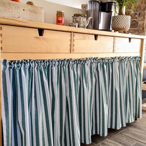 May include: A wooden cabinet with three drawers and a green and white striped fabric valance. The cabinet is topped with a coffee maker, a teapot, and jars of spices.