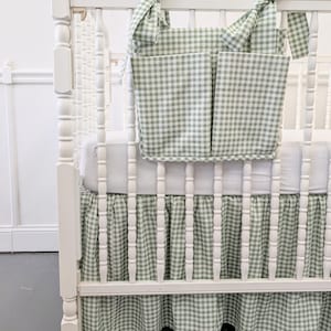 Könnte beinhalten: Ein weißes Babybett mit grün-weißem Vichy-Muster. Das Babybett verfügt über einen passenden Bettrand und einen hängenden Organizer mit zwei Taschen. Der Organizer ist mit Schleifen am Babybett befestigt.