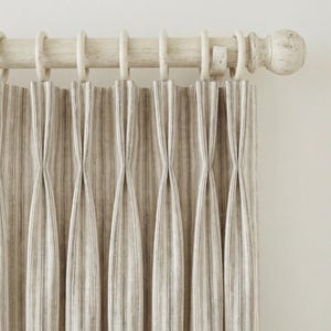 May include: Beige and gray striped curtains hanging from a distressed white wooden rod with rings. The curtains have a pleated design, creating vertical lines. The rod has a decorative finial on the end.