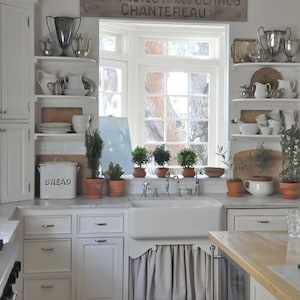 May include: A white kitchen with a farmhouse sink, white cabinets, and open shelving. The shelves are decorated with vintage teapots, cups, and other kitchenware. A wooden sign above the window reads "FARAPLUIES Produits Centres CHANTEREAU".