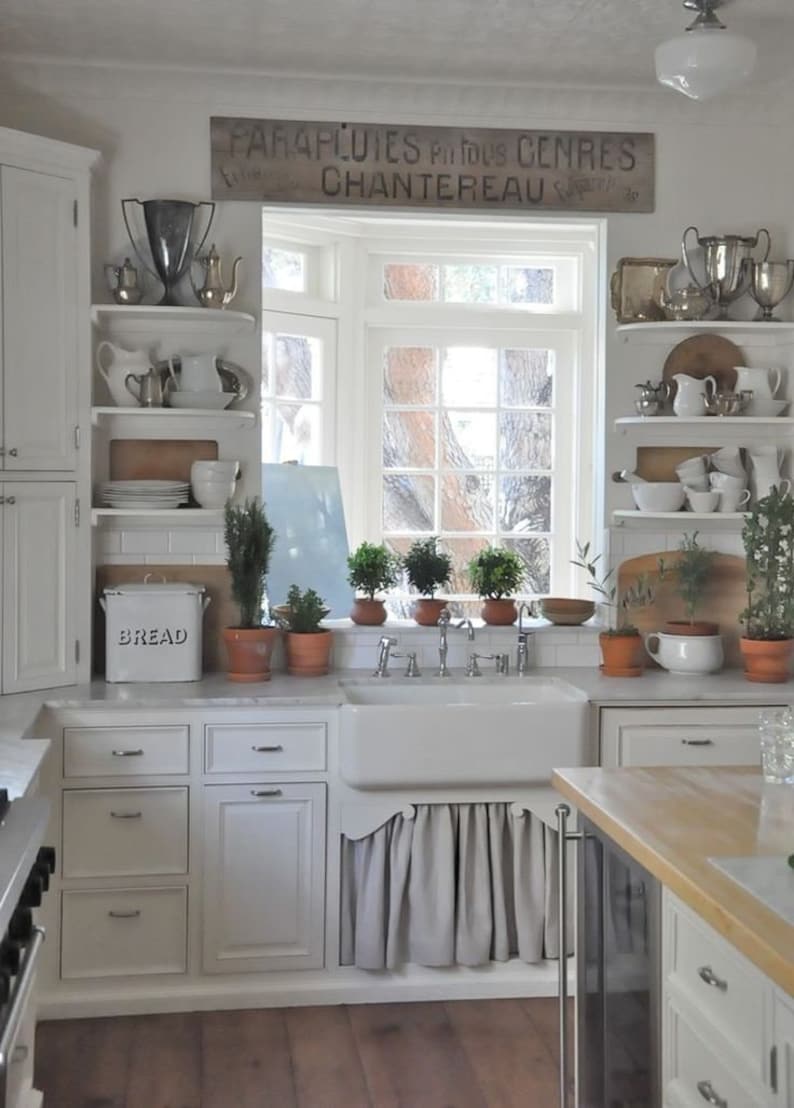 May include: A white kitchen with a farmhouse sink, white cabinets, and open shelving. The shelves are decorated with vintage teapots, cups, and other kitchenware. A wooden sign above the window reads "FARAPLUIES Produits Centres CHANTEREAU".