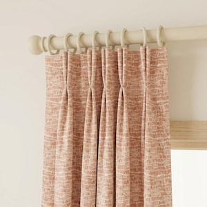 Puede incluir: Cortinas plisadas con un patrón repetitivo de líneas verticales finas en un tono marrón rojizo cálido sobre un fondo crema. Las cortinas cuelgan de una barra de madera de color claro con anillos a juego.