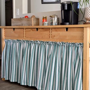 May include: A wooden cabinet with three drawers and a green and white striped fabric valance. The cabinet is topped with a coffee maker, jars, and a teapot.