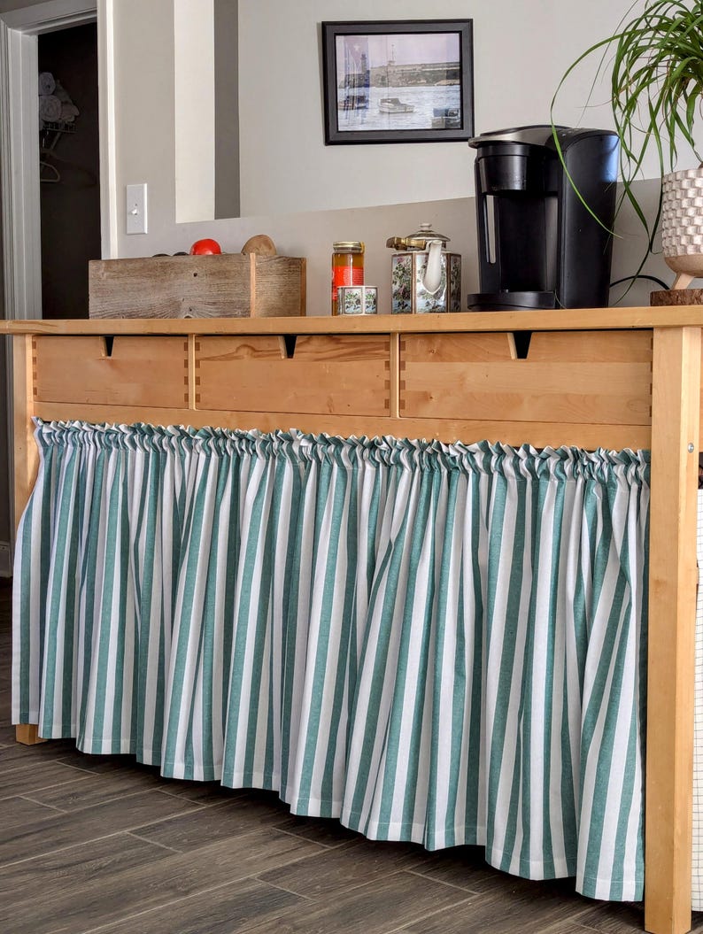 May include: A wooden cabinet with three drawers and a green and white striped fabric valance. The cabinet is topped with a coffee maker, jars, and a teapot.