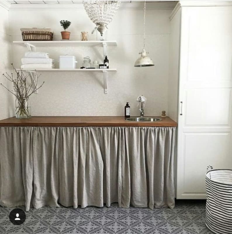May include: A white laundry room with a wooden countertop and a sink. The countertop is covered with a light gray linen curtain. The room has white shelves with towels and a basket on the top shelf. A white cabinet is on the right side of the image. The floor is tiled with a black and white pattern.