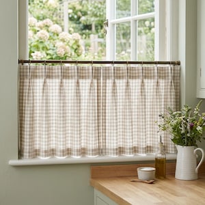 Op de afbeelding: Een paar beige en witte gingham café-gordijnen met plooien, hangend aan een donkere metalen roede met ringen. De gordijnen bedekken de onderste helft van een raam en zijn voorzien van een klassiek ruitpatroon en een gestructureerde bovenkant.