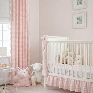 Könnte beinhalten: Ein Kinderzimmer mit einem weißen Kinderbett, rosa Vichy-Bettwäsche und einem passenden Bettrand. Rosafarbene Vorhänge mit weißer Pompon-Borte umrahmen ein Fenster. Plüschtiere, darunter ein Schwein und ein Lamm, sitzen auf dem Boden. Zwei gerahmte Bilder hängen an der Wand.